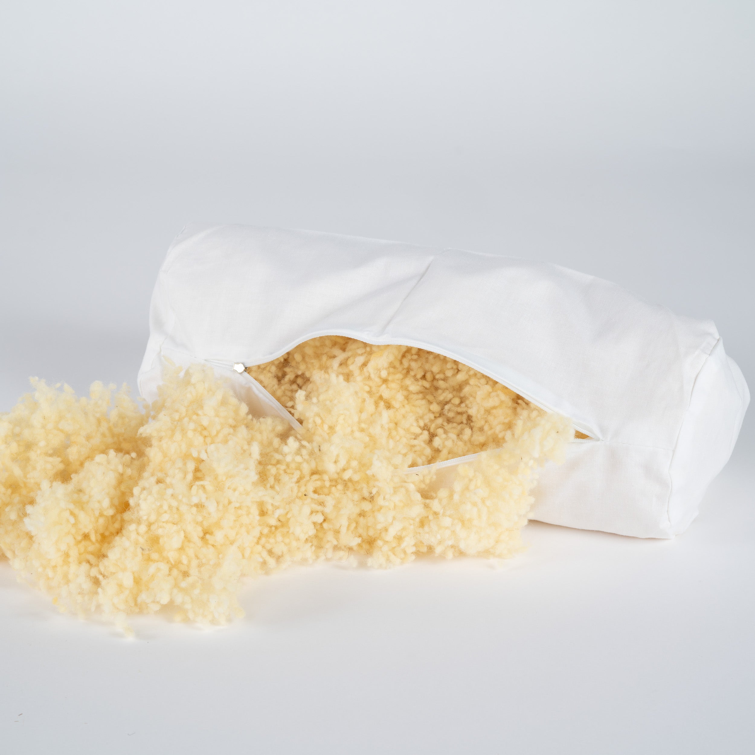 Weißer Nackenrollenbezug mit Reißverschluss, aus dem fluffige Füllung aus naturfarbenen Schurwollkügelchen quillt